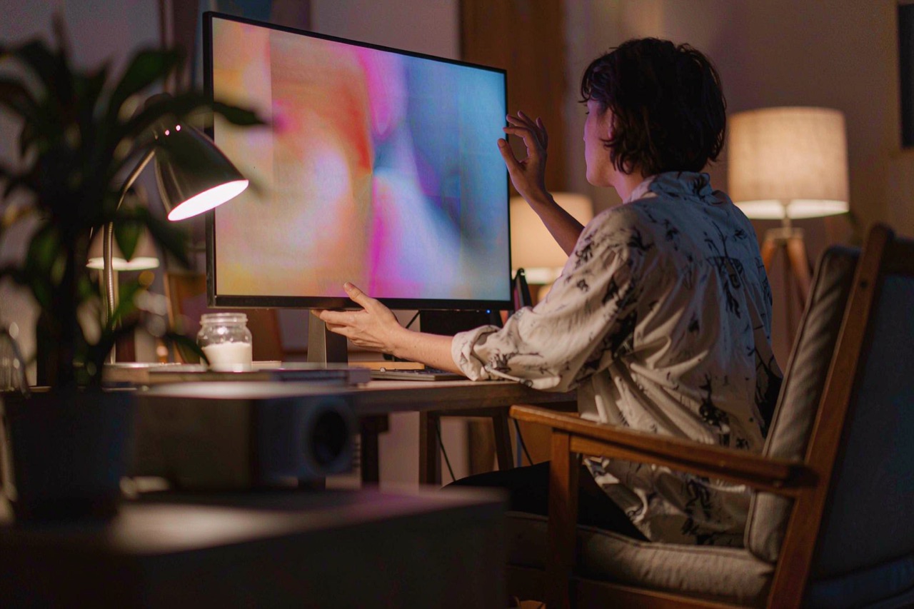 A person adjusting a large monitor in a cozy evening home office with warm lamp light