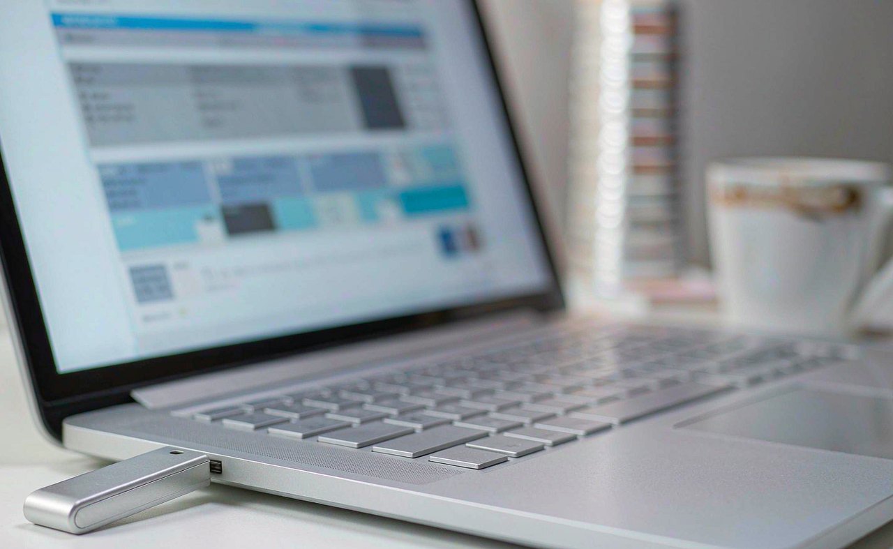 A USB flash drive plugged into the side of a laptop displaying a blue desktop, with a notebook and coffee mug on a tidy desk