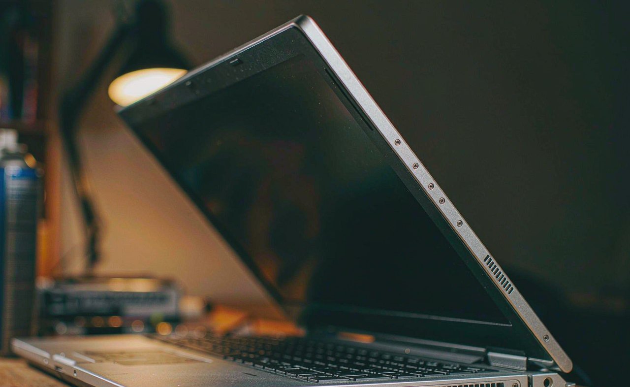 A laptop with its side cooling vents and a screwdriver on a wooden desk beside a can of compressed air, warm desk lamp glow, photorealistic