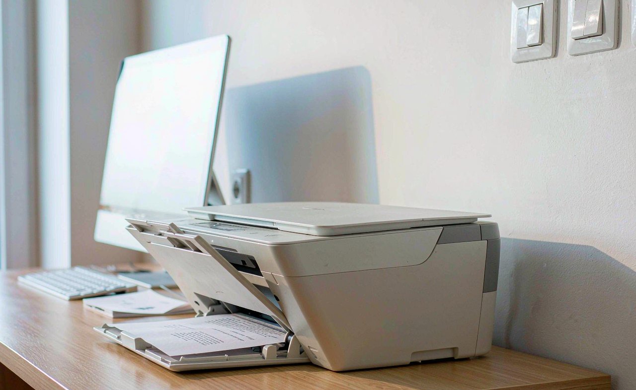 A compact inkjet printer on a light wooden desk with a printed page emerging from its tray, beside a laptop in a bright home office