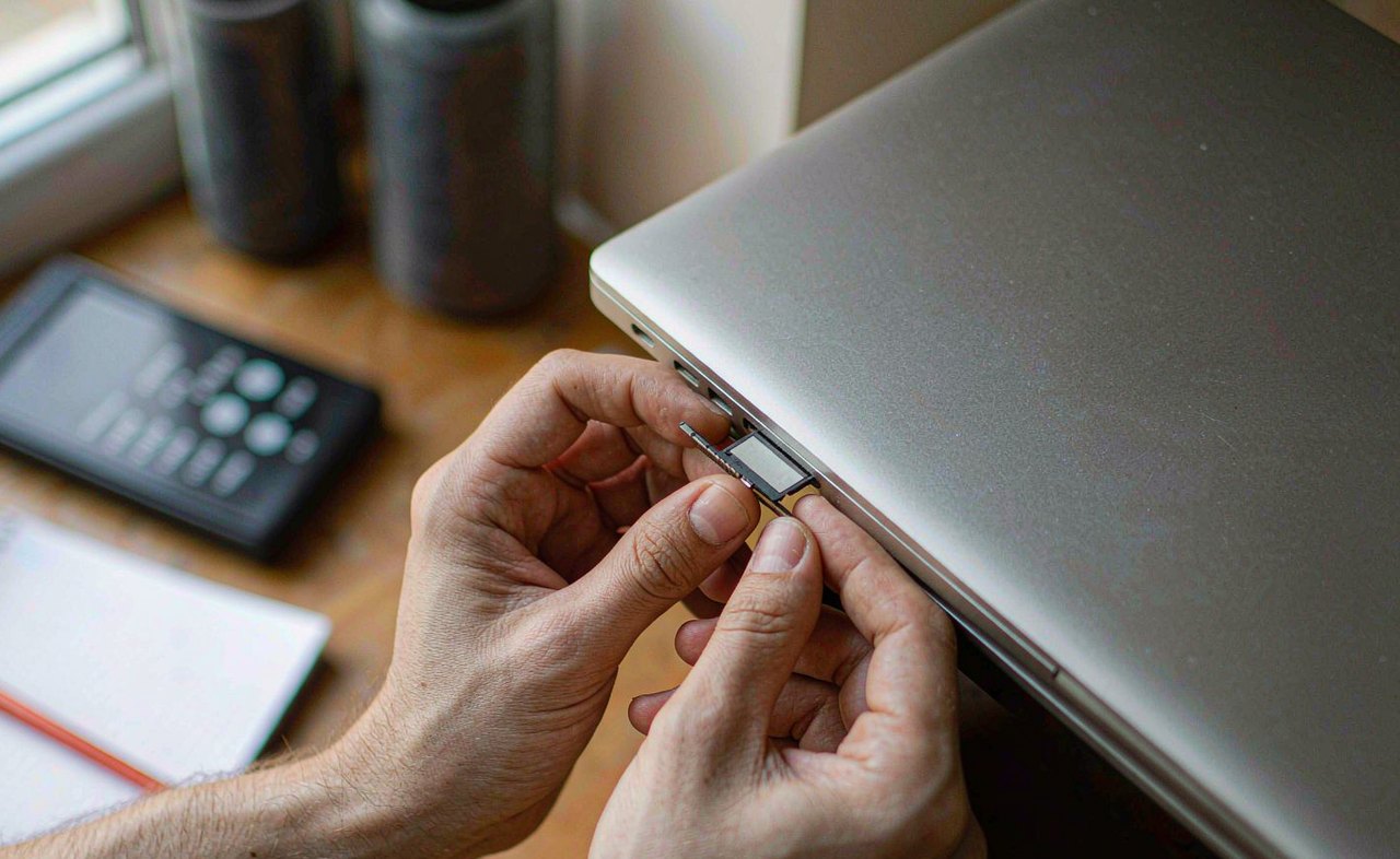 Close-up of hands inserting an SD card into the side of a silver laptop on a wooden desk in warm afternoon light