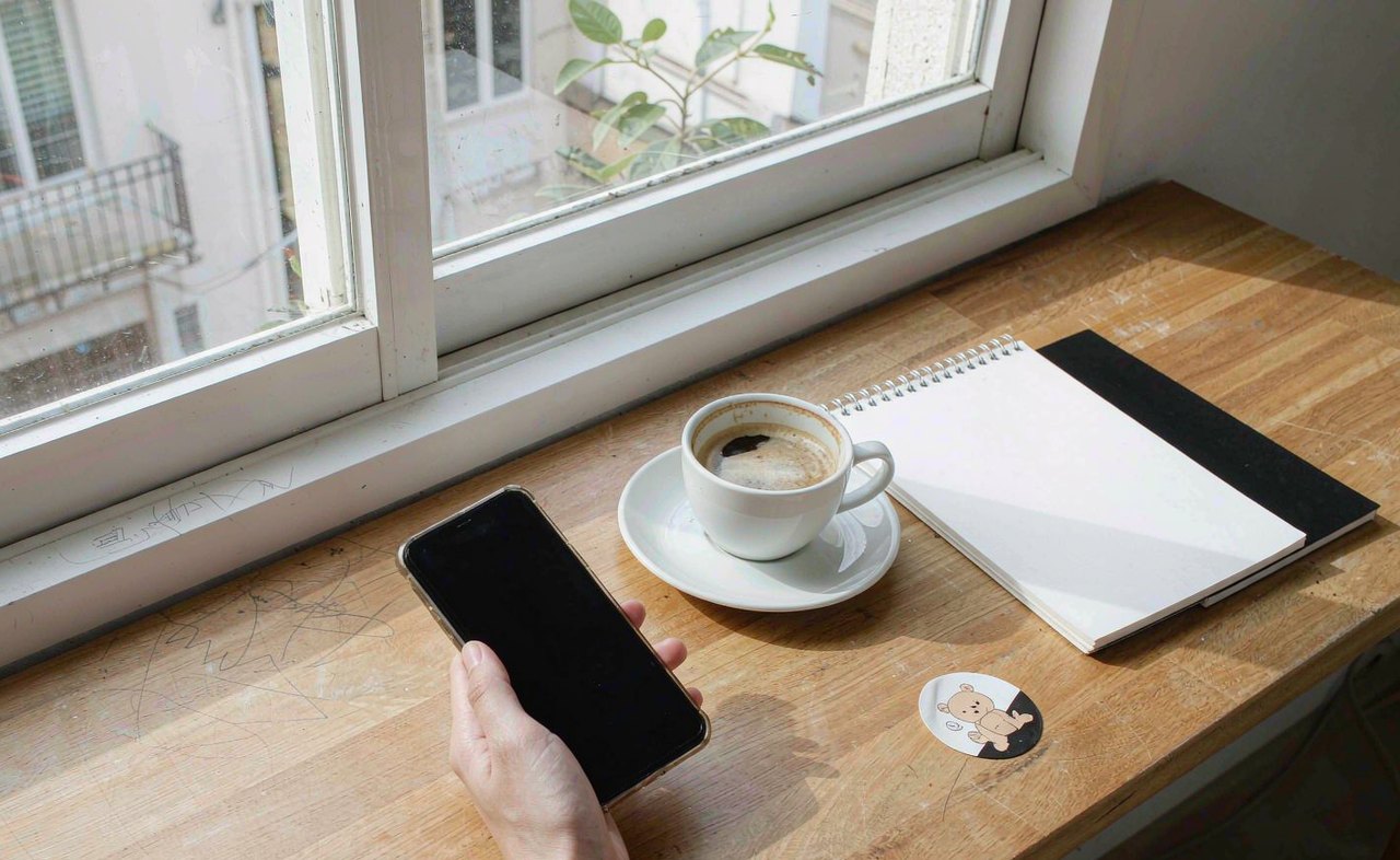 A hand picking up an iPhone from a tidy wooden desk beside a notebook and a coffee cup, soft morning window light, realistic photo style