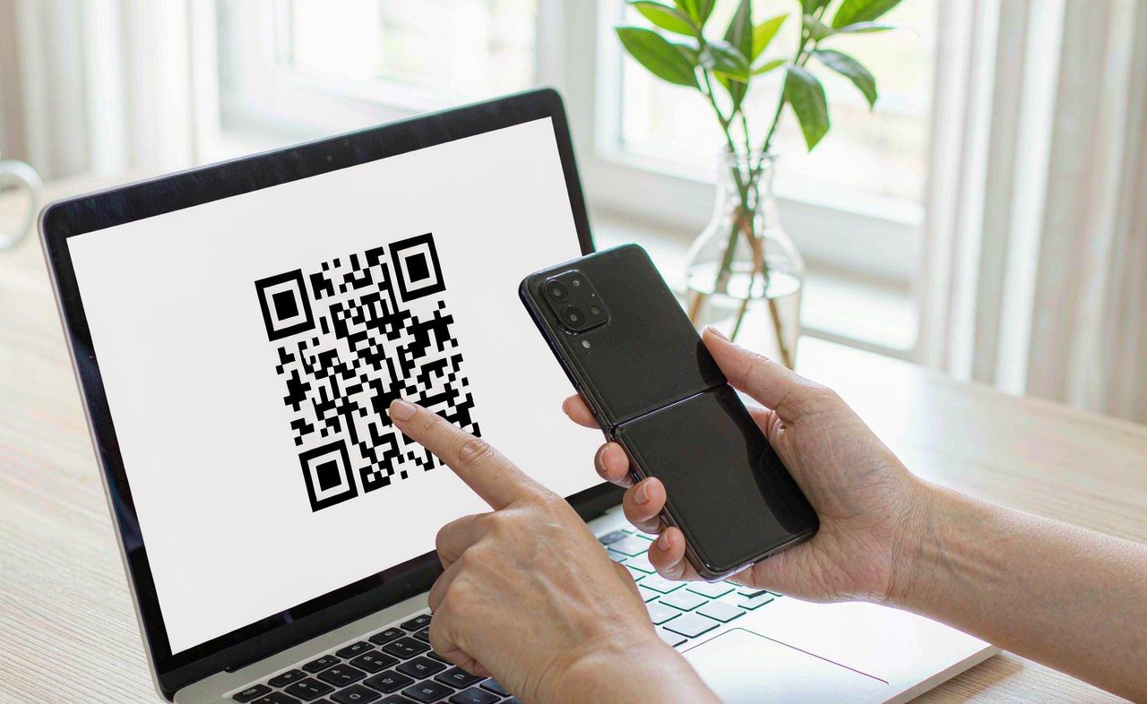 Hands holding a smartphone scanning a QR code shown on a laptop screen in a bright home office, morning window light, photorealistic