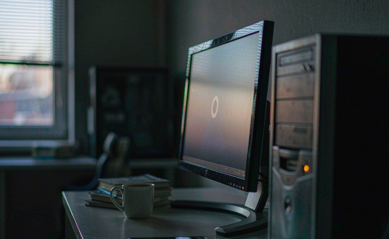 A desktop PC tower and monitor glowing in a dim room at dawn, power light on